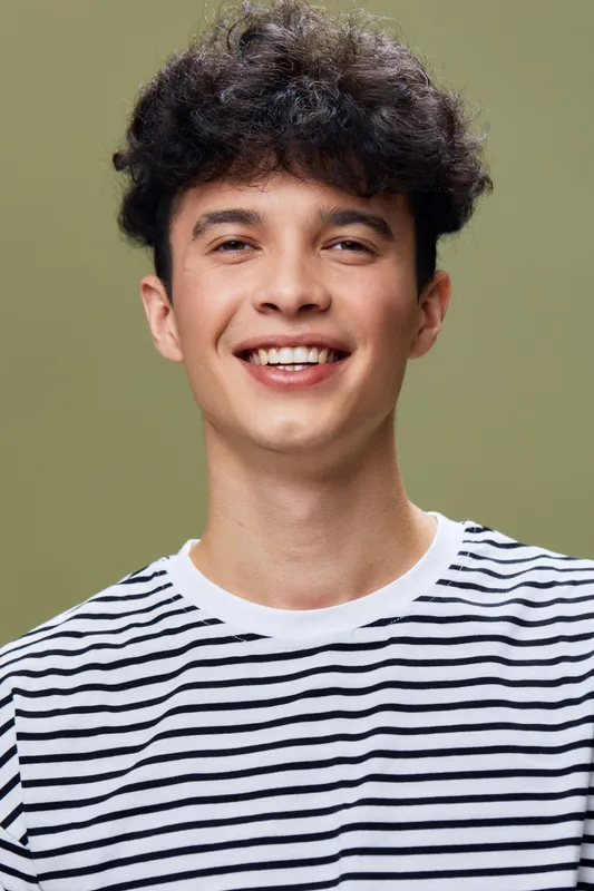smiling man in striped shirt with curly hair, posing against olive green background, cheerful young male expressing happiness and confidence with natural light
