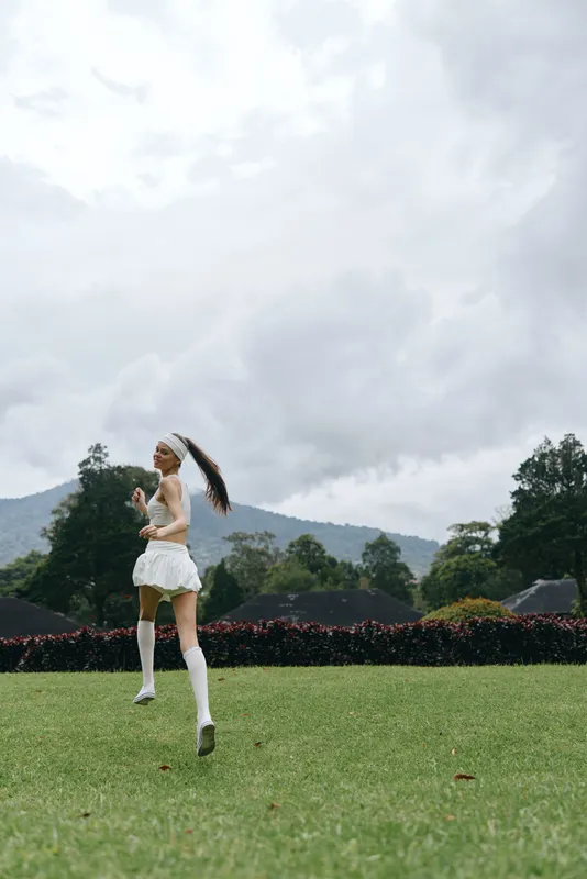 Young female runner in a white outfit joyfully exercising outdoors on a grassy field with a mountainous backdrop, emphasizing a healthy lifestyle and fitness concept