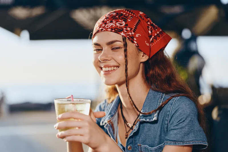 smiling woman with red bandana enjoying drink outdoors on sunny day with blurred background and casual denim shirt, happiness and relaxation captured naturally