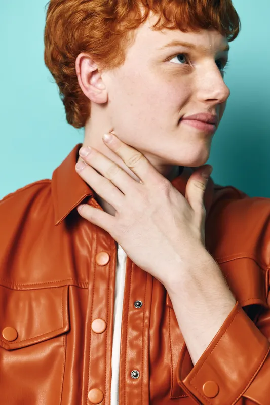 Young man with red hair wearing brown leather jacket looks sideways with hand on neck against light blue background in studio. Portrait of casual fashion model with confident expression.