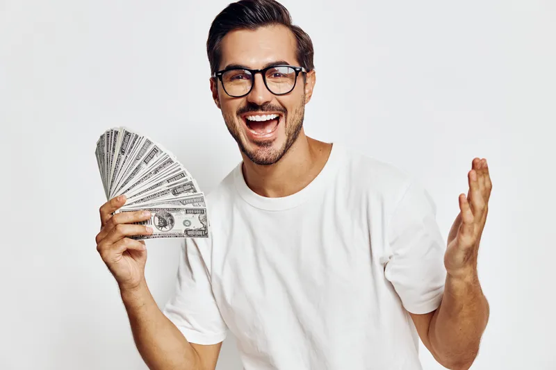 Full body smiling happy cheerful young man wear white t-shirt look camera hold in hand stack of money isolated on plain solid light background studio portrait. Lifestyle concept