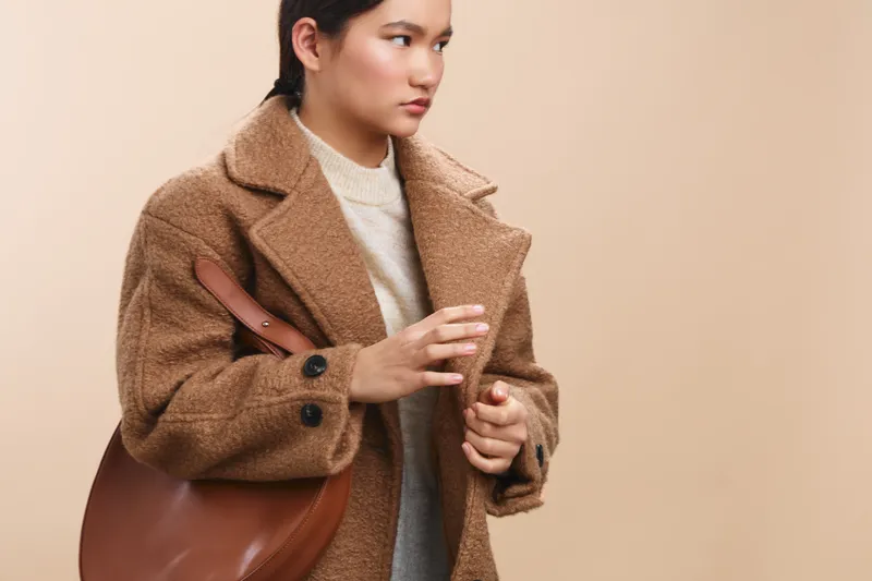 asian woman wearing a warm brown coat and carrying a leather bag, standing against a neutral background. She gazes to the side with a calm expression, conveying autumn style and quiet confidence.