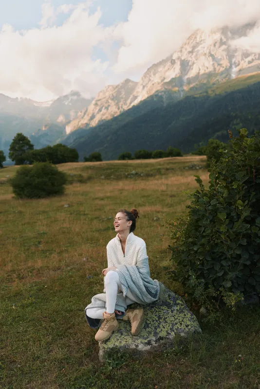 A young woman in casual clothes sits on a rock in a mountain meadow laughing joyfully while wrapped in a blanket during a sunny day in nature.