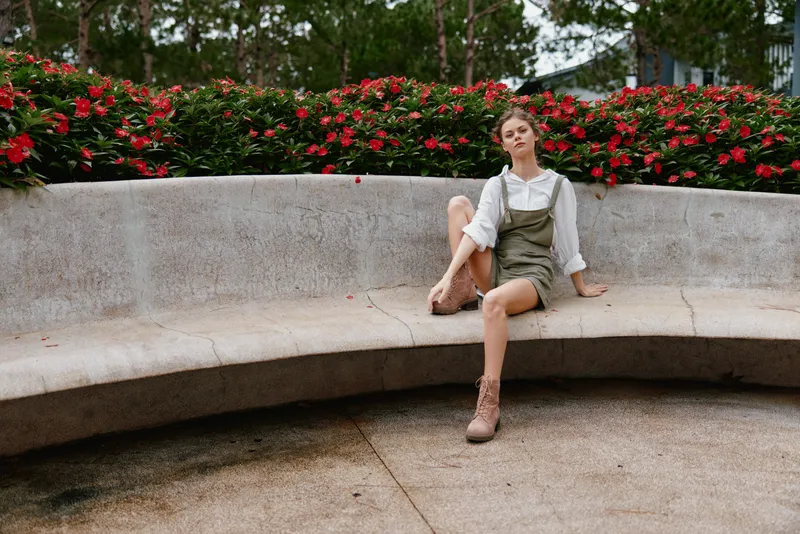 Stylish young woman in a casual summer outfit sitting on a curved bench surrounded by vibrant red flowers, expressing relaxed confidence and an outdoor lifestyle