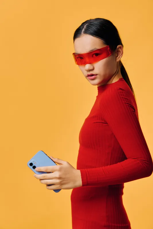 Young woman in red dress and futuristic red glasses holding blue smartphone against orange background looking at camera. Studio portrait with modern fashion and technology style.