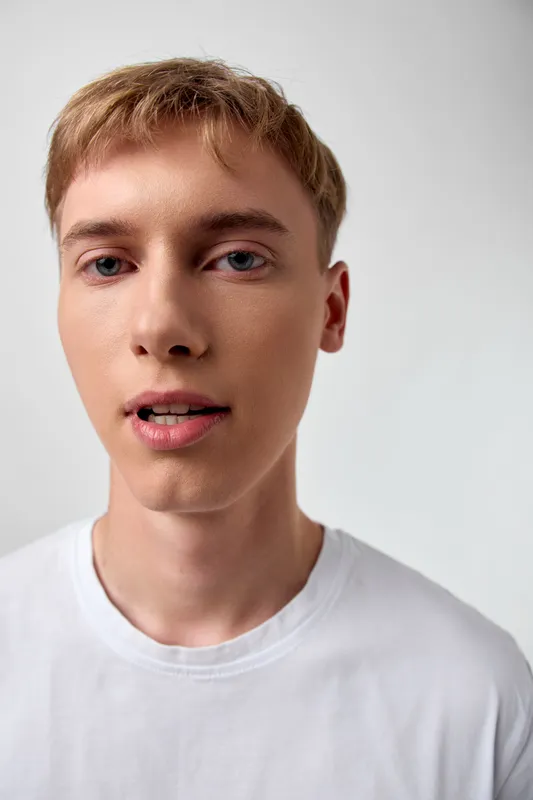 Close-up of a young man with a calm expression wearing a white t-shirt, set against a neutral background with studio lighting for a clean, minimalist image suitable for profile and fashion use