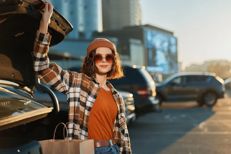 woman shopping by car trunk at sunset in urban parking, portrait of beanie style with sunglasses and bag, candid lifestyle authenticity and golden hour glow, mindful living mood