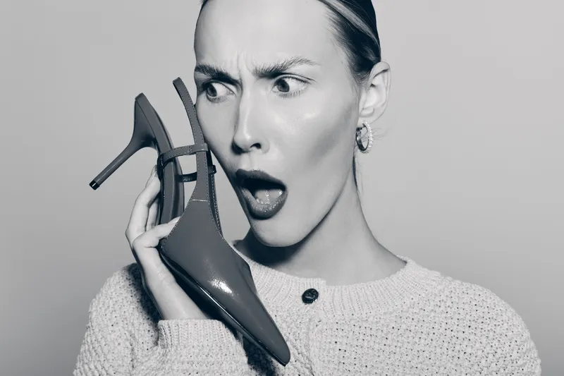 Surprised Caucasian woman with light skin holds high heel shoe near face, expressing shock and disbelief. Studio portrait in monochrome with casual sweater and earrings.