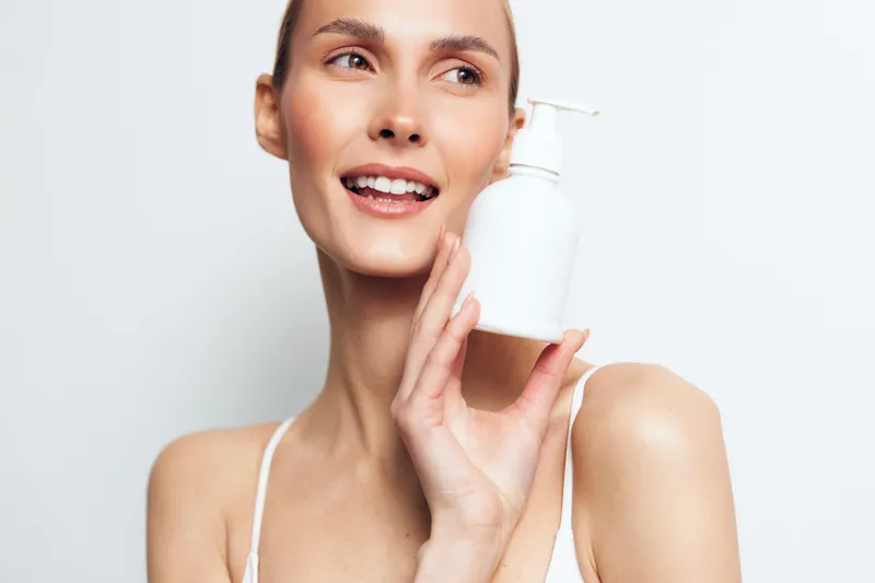 A smiling young woman in white holds a white cosmetic bottle near her face in a bright studio. The image represents skincare, beauty, and personal care concepts for health and wellness.