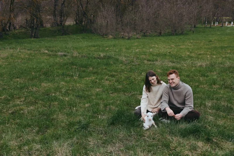 Two people sit on grass in a park with a small white dog, enjoying a relaxed moment together, showcasing affection and lifestyle through candid outdoor bonding and cheerful companionship