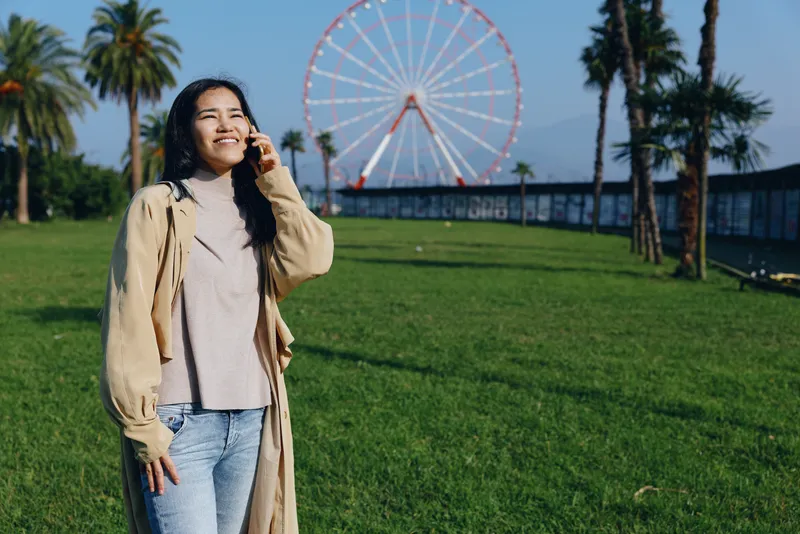 Woman in an outdoor park, casual cardigan and jeans, smiling beside a ferris wheel, sunlit grass and blue sky, leisure travel mood, cheerful and relaxed, vibrant urban nature backdrop