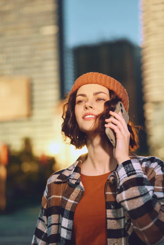 Woman phone urban golden hour beanie candid authenticity lifestyle smiling portrait of a young woman on a city street, warm sunlight glow, mindful living and emotional storytelling in a modern moment