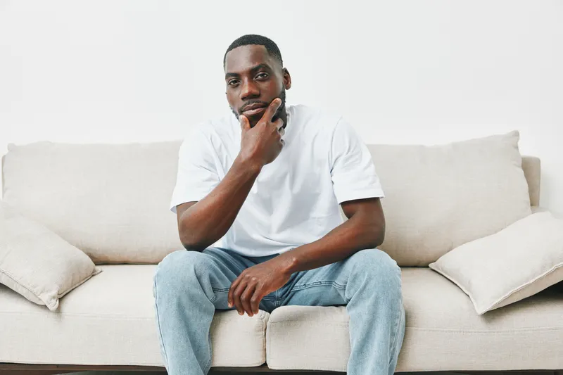 Thoughtful young man in casual white t shirt and jeans, sitting on a light colored sofa, reflecting on life and emotions in a minimalist setting