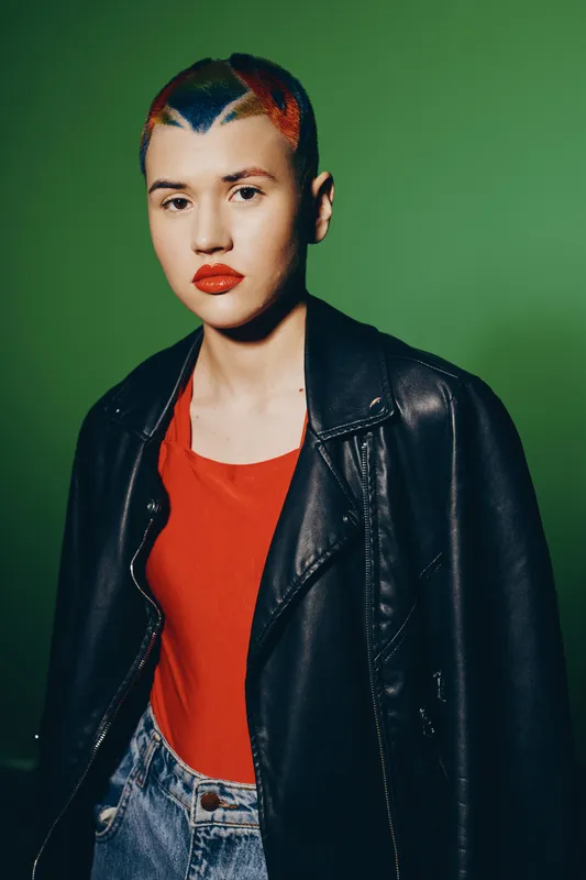 young woman with short dyed hair wearing a black leather jacket and red top standing against a green background, showcasing modern style and bold makeup.