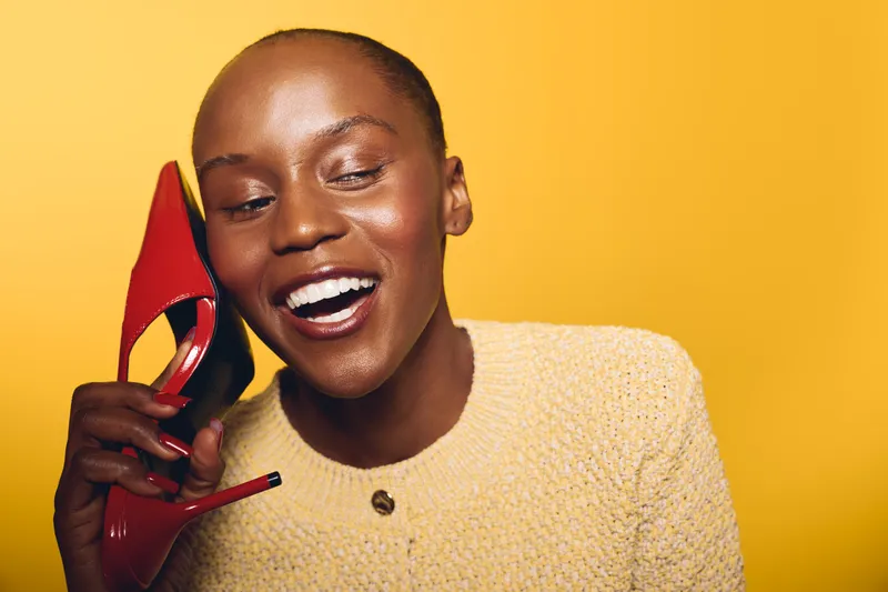 Happy African woman with dark skin smiling and holding red high heel shoe near face on yellow background. Concept of fashion, beauty, lifestyle, and joyful expression.