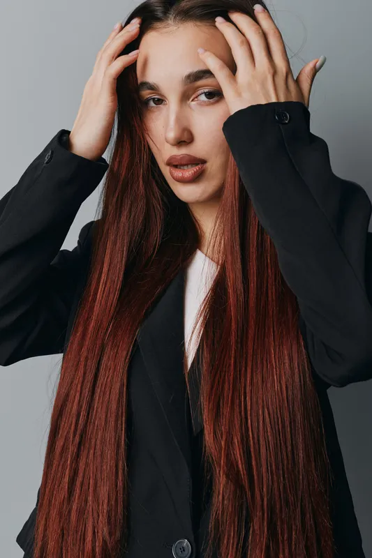 Young woman with long straight hair wearing black jacket posing with hands on head against gray background. Fashion model with natural makeup and serious expression in studio.