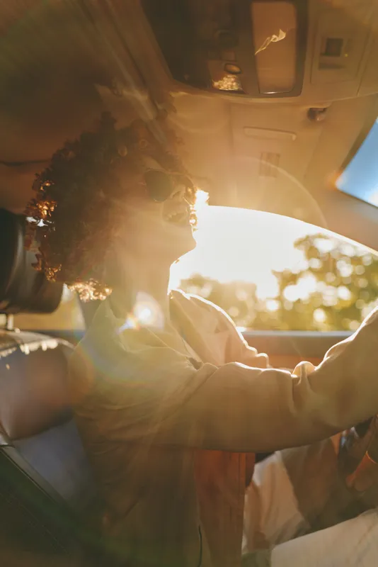 A woman driver sits behind the wheel in a vintage car, hands on the steering, sunlit window glow, retro film color. A casual lifestyle moment that hints at road trips, freedom, and warm nostalgia.
