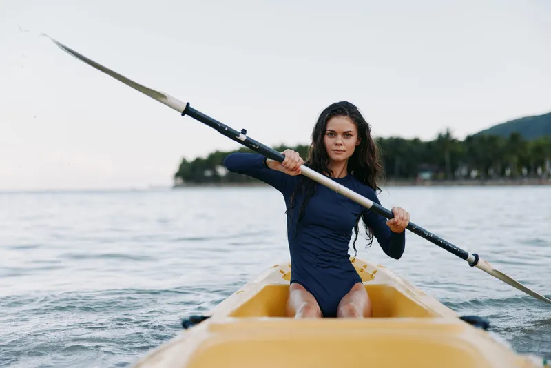 Woman kayaking on calm water wearing navy blue swimsuit, focused and active, surrounded by peaceful nature with soft light and distant trees.