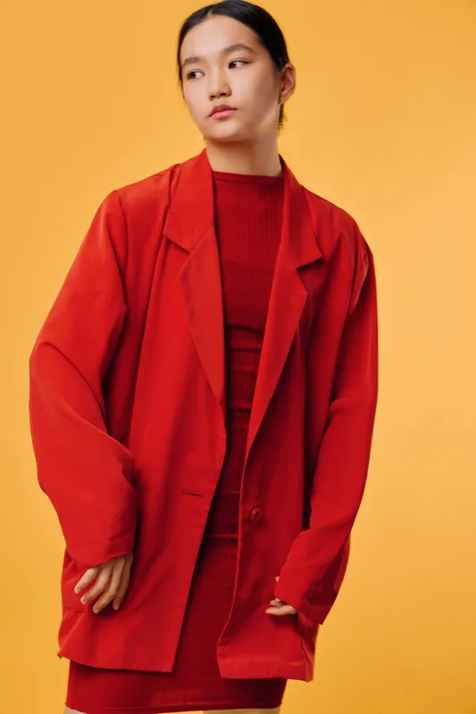 Woman wearing oversized red blazer and matching red dress standing against orange background with minimal expression. Fashion model posing with straight hair and natural makeup.