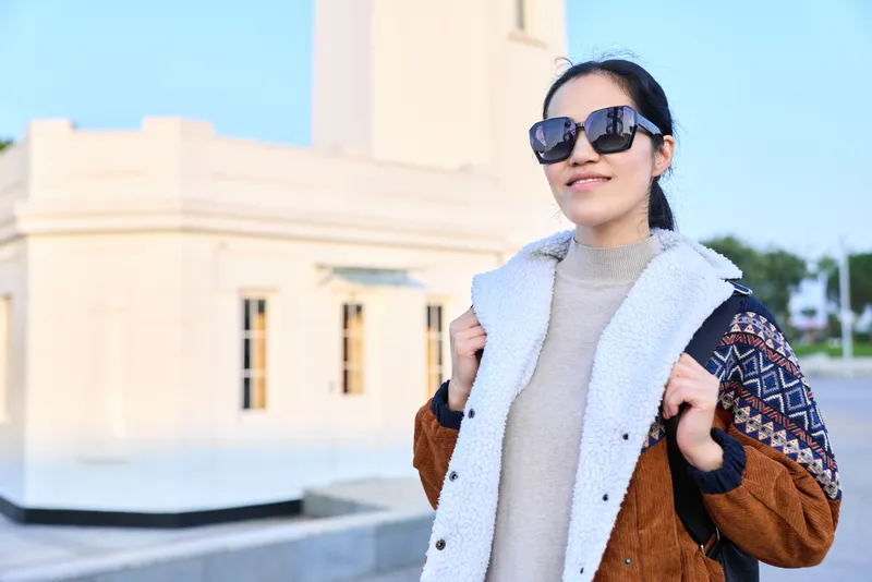 Fashion, outdoor, sunglasses, coat, jacket style showcased by a chic traveler in an urban setting on a sunny day with a cityscape backdrop and confident casual attire