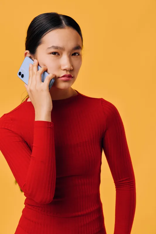 Young woman in red long sleeve shirt holding smartphone to ear with serious expression on orange background. Portrait of female talking on mobile phone with focused look.