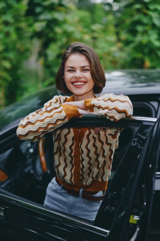 Smiling woman leans from a car window, wearing a striped knit sweater, enjoying a candid outdoor moment.