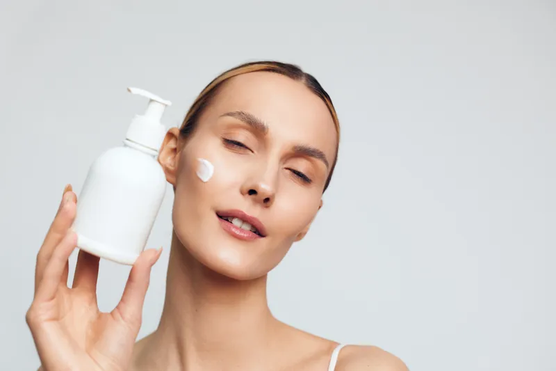 A young woman with brunette hair applies cream from a pump bottle to her cheek, capturing a focused skincare moment that conveys calm relaxation and self care.