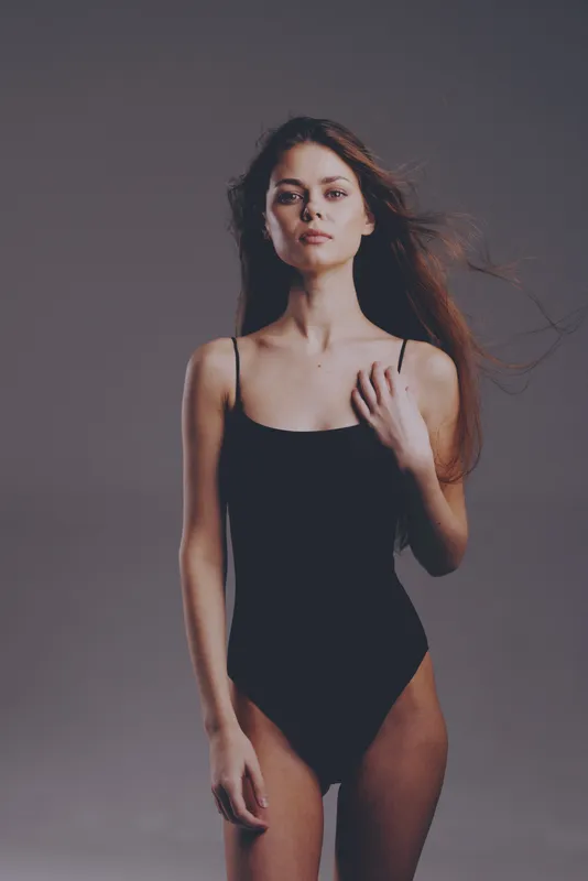 Young woman in black bodysuit posing with neutral expression against gray background
