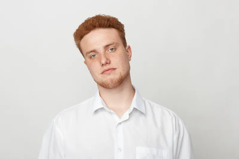 Young man with red hair and beard wearing white shirt looks at camera with neutral expression on light background. Concept of casual style, portrait, and natural appearance.