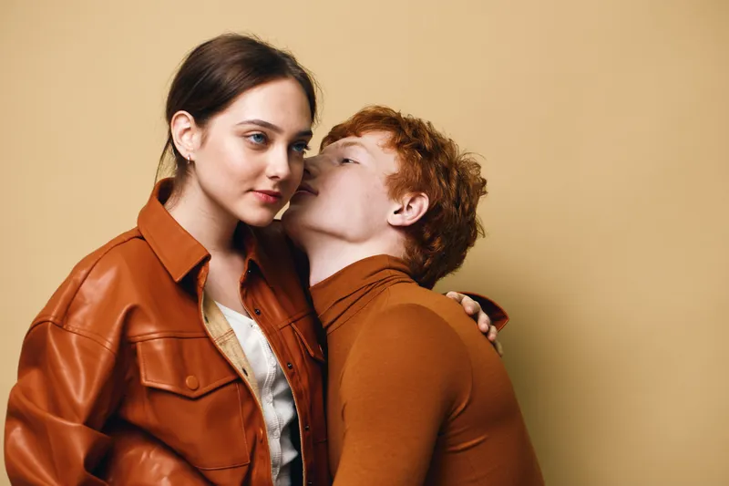 Couple with red hair and brown jacket in loving embrace against beige background. Intimate moment, affection and connection with casual style and soft lighting.