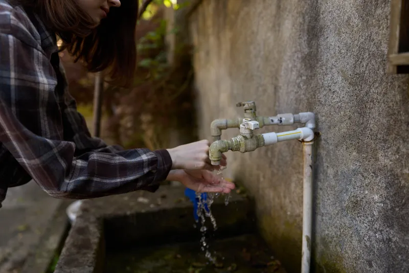 Young woman in a plaid shirt washes her hands at an outdoor faucet, water splashes and drips, sunlight highlights a weathered wall in a quiet backyard