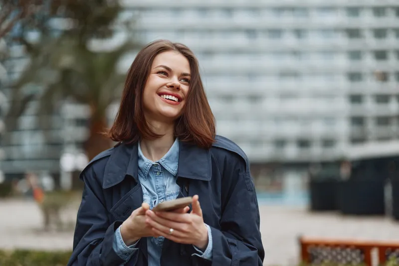Young woman with brunette hair smiles while holding a smartphone in a denim jacket and light blue shirt, outdoors in a bright urban setting, radiating warmth and friendliness.