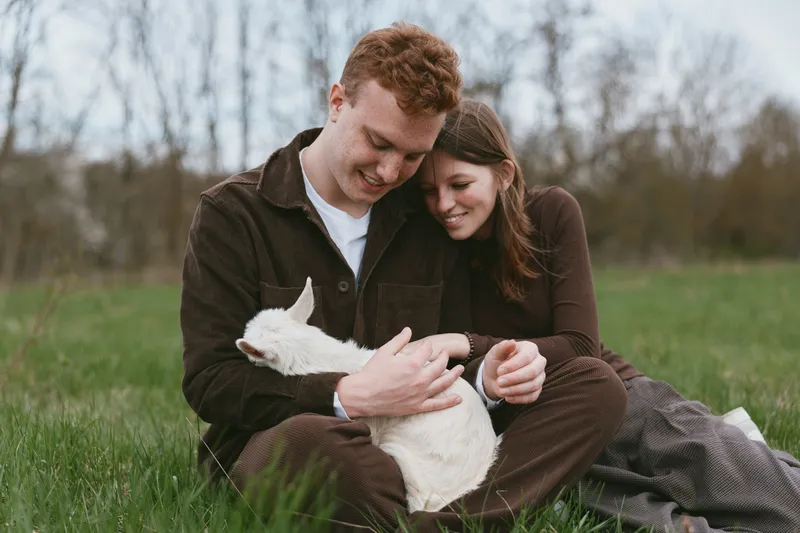 A joyful couple sits on grass in a sunlit field, gently holding a small goat kid between them. This lifestyle moment highlights affection, bonding, and rustic countryside joy.