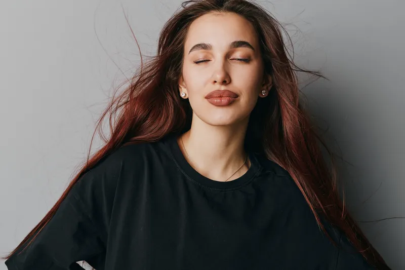 Woman with long hair and closed eyes wearing black shirt posing against gray background for beauty, relaxation, and calm expression in studio portrait with natural makeup and earrings.
