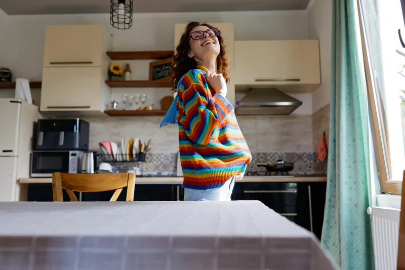 A woman wearing a colorful striped sweater stands in a bright kitchen, capturing a confident, casual home lifestyle moment for everyday living and domestic aesthetics.
