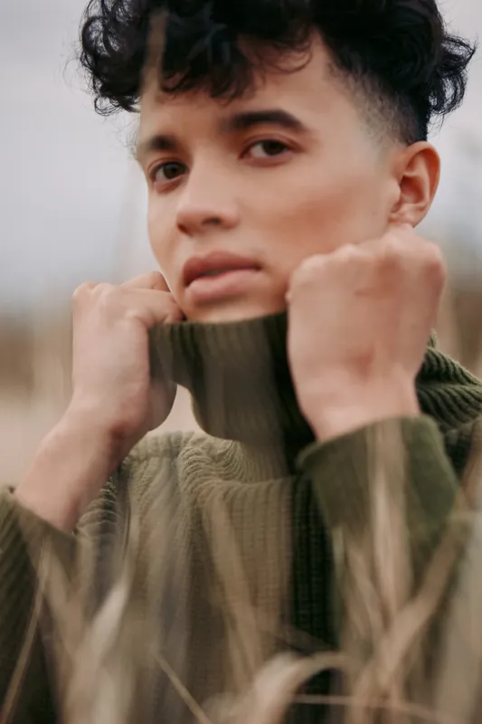 Close-up portrait of a young person in a green turtleneck among tall grass, natural gaze and subtle expression conveying an authentic, credible mood and an honest contemporary portrait.