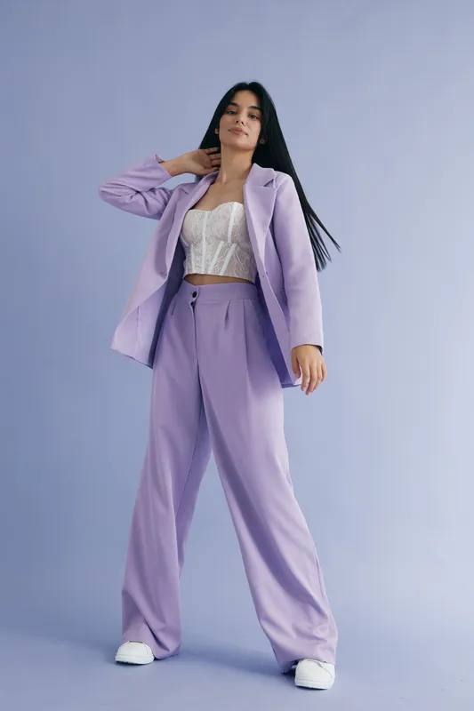 Woman posing in lilac pantsuit with white lace top against a pastel purple background. Stylish, confident, modern fashion outfit for young adult.