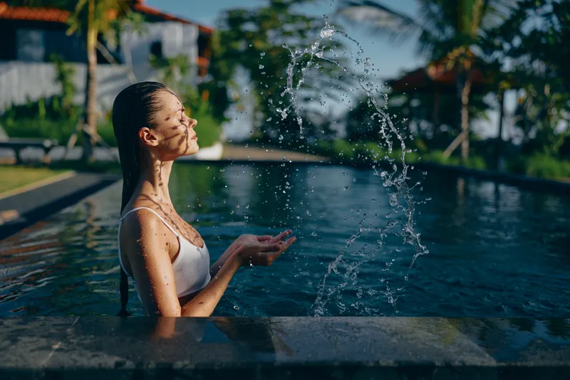 Young woman in white swimsuit playing with water in outdoor pool during sunny day. Relaxed lifestyle concept with natural light and tropical background creating calm mood.