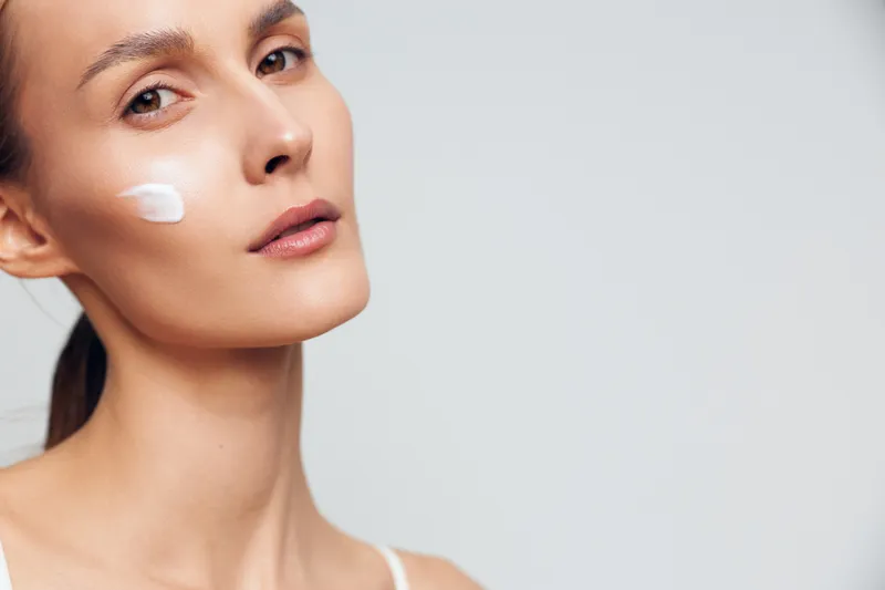 Young woman with clear skin applies moisturizing cream on cheek looks at camera with calm expression isolated on light background skincare beauty concept.