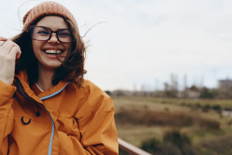 Smiling woman in orange jacket and knit beanie outdoors, wearing glasses, enjoying fresh air, lifestyle portrait with soft daylight and distant fields in background, casual winter mood today