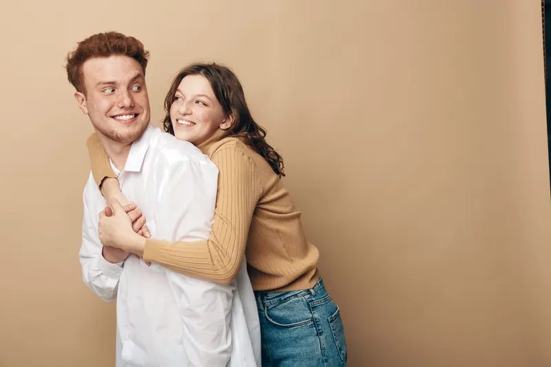 Happy young couple embraces and smiles looking away on beige background. Casual clothing, positive mood, love and friendship concept with natural light and warm tones.