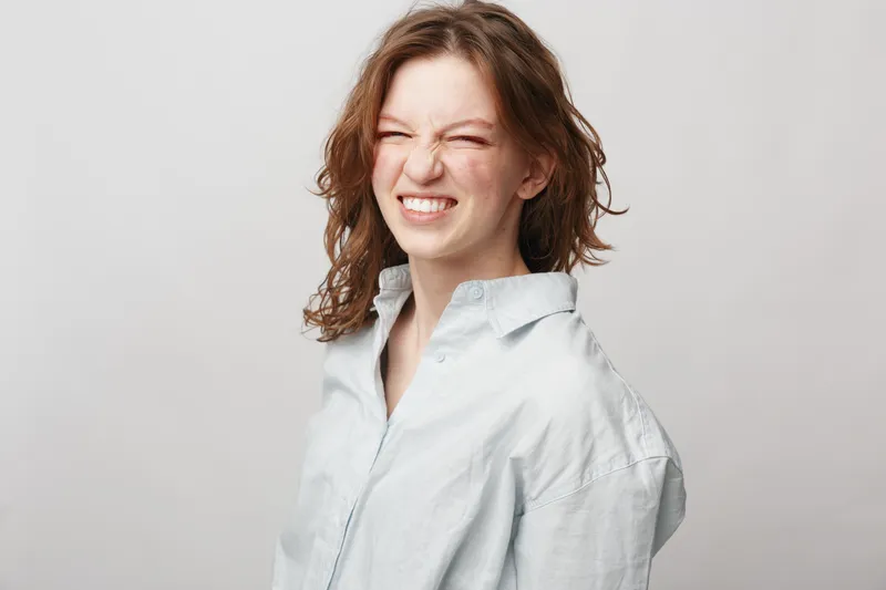 Young woman with curly hair wearing a light shirt makes a funny grimace with eyes closed. Studio portrait on white background. Concept of emotions, humor, and natural expression.