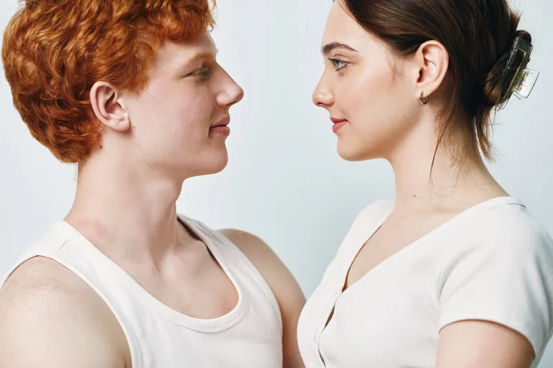 Man and woman with red hair and dark hair looking at each other with calm expressions on white background. Closeup portrait of young adult people facing one another.