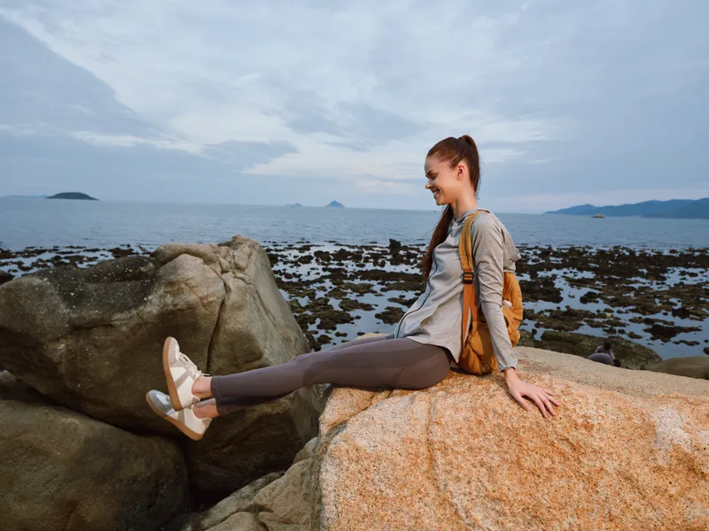 Young woman enjoying nature on rocks by the sea with a relaxed expression, wearing athletic clothing in soft colors suitable for outdoor activities