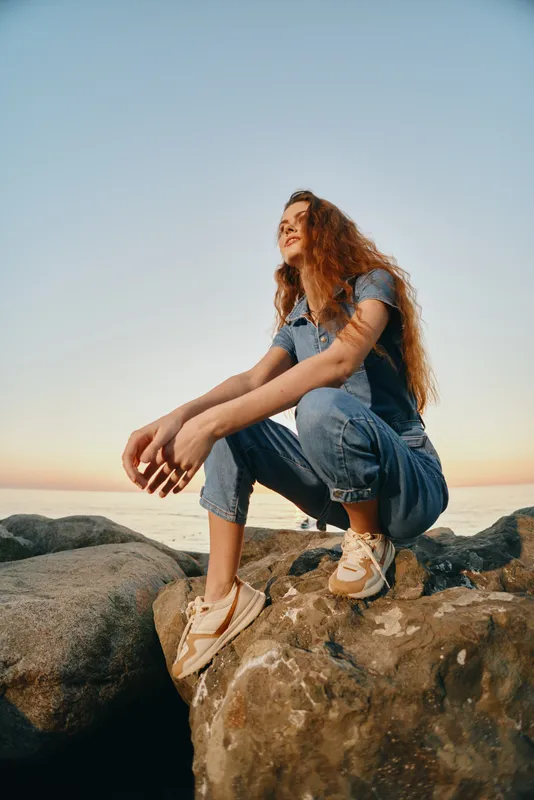 young woman sitting on rocks during sunset, wearing casual denim outfit and sneakers, enjoying outdoor nature with a relaxed and thoughtful mood