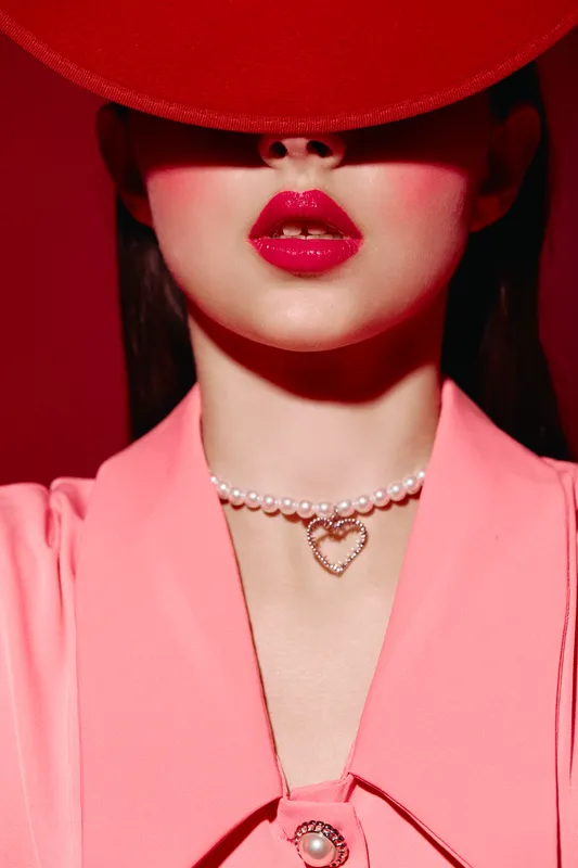 Close-up of a woman with glossy red lipstick, wearing a pink outfit and pearl necklace with heart pendant under dramatic shadow from a wide red hat. Beauty fashion concept.