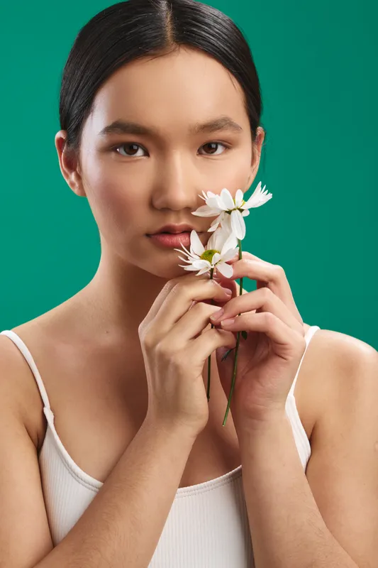 Asian woman holds white flowers near her face, a soft expression and calm gaze in a studio portrait against a teal background, creating a serene, elegant mood for beauty, wellness, and lifestyle