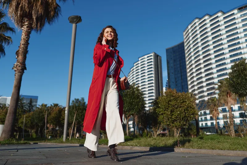 Brightly dressed woman in a red coat walks through a sunny urban park with tall buildings behind, exuding confidence and a relaxed vibe.