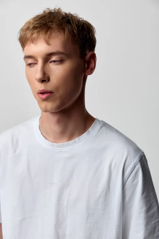 Calm man in a white t shirt stands in a studio with soft lighting, a relaxed pose and distant gaze, creating a minimalist mood for fashion, lifestyle and wellness projects