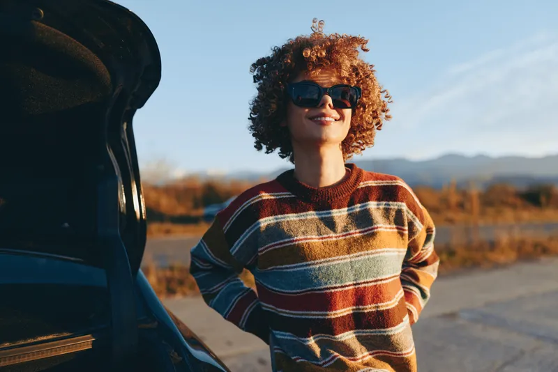 Woman with curly hair smiling outdoors, wearing striped sweater, standing near car on road, sunlight golden hour, relaxed lifestyle mood, soft breeze, warm natural colors, distant hills